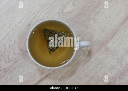 Top-down-Perspektive des grünen Teebeutels, der in goldener Flüssigkeit in einer weißen Tasse auf einem hellen Holztisch infundiert wird, saubere Zusammensetzung Stockfoto