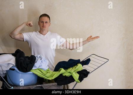 Ein energiegeladener junger Mann in weißem T-Shirt posiert mit einem Arm gebeugt und einem anderen in der Nähe eines Wäschestücks voller Wäsche und blauem Korb Stockfoto