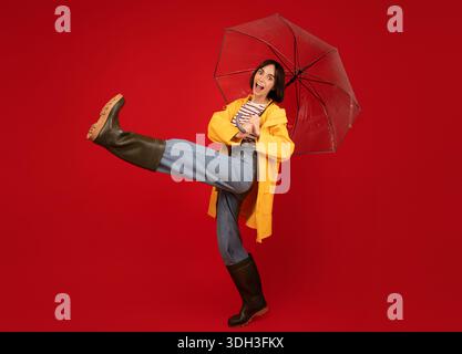 Positive Dame in guter Laune mit Stiefeln und gelber Regenjacke, die unter einem transparenten Regenschirm über rotem Hintergrund läuft Stockfoto
