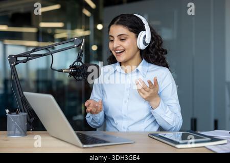 Junge Frau, die Kopfhörer trägt und mit animierten Gesten in ein Mikrofon spricht, während sie einen Podcast aufnimmt oder online Live-Streaming aus einem modernen Bürostudio streamt Stockfoto