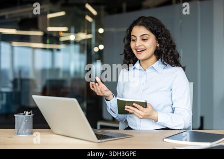 Junge Geschäftsfrau mit dunklen lockigen Haaren, die an einem Schreibtisch sitzt. Aktive Teilnahme an einem Videoanruf auf ihrem Laptop. Halten eines Notizbuchs. Und Gesten während der Kommunikation von einem modernen Arbeitsplatz aus Stockfoto