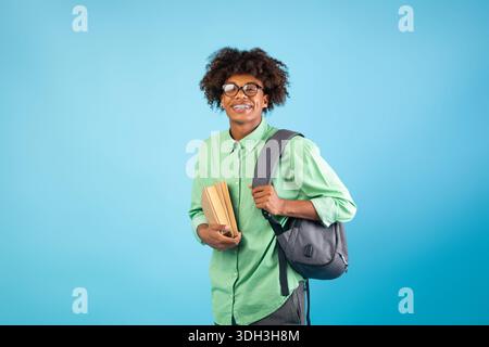 Fröhlicher schwarzer Student mit Rucksack und Büchern, die über dem blauen Studiohintergrund die Kamera anlächeln Stockfoto
