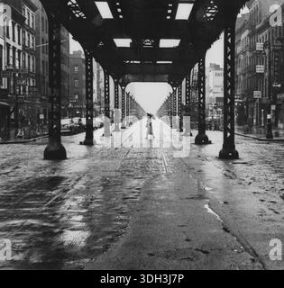 Elevated Train Tracks NYC – das Foto zeigt eine Frau mit einem Regenschirm, die die Straße überquert, eingerahmt von den Säulen einer Unterbauüberführung. Kreuzweg im Bild ist möglicherweise E 41st St., 1956 – Rizzuto, Angelo, 1906–1967, Fotograf. Stockfoto