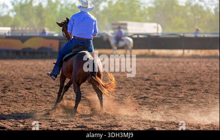 Ein Blick von hinten auf einen Cowboy mit weißem Hut und blauem Hemd, der auf seinem Pferd sitzt und während eines Rodeos über eine staubige Arena blickt. Stockfoto