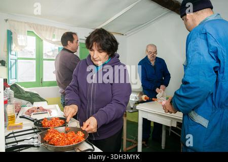 Eine ältere Frau kocht Chorizo ​​meat mit ihrer Familie, die in der Nähe des alten Hauses während einer Pause vom Tag der Schweineschlachtung interagiert. Stockfoto