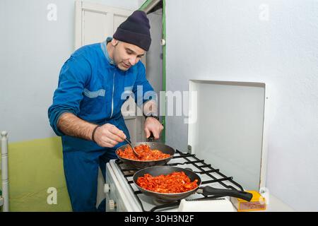 Der fröhliche Mann in blauen Overalls rührt die gewürzte Schweinefleisch-Mischung auf, während sie auf dem alten Dorfgasofen kocht. Stockfoto
