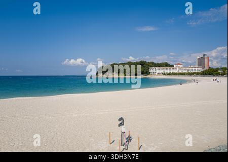 Shirarahama, Japan - 25. Oktober 2023: Ein weiträumiger weißer Sandstrand mit türkisfarbenem Wasser, ein paar Menschen entlang der Küste und mehrere moderne Gebäude und Hotels, die im Hintergrund in der Nähe eines bewaldeten Gebiets unter einem klaren blauen Himmel zu sehen sind. Stockfoto