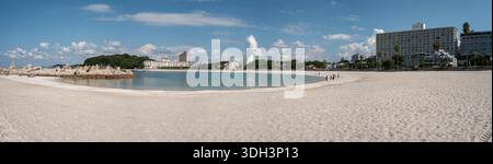 Shirarahama, Japan - 25. Oktober 2023: Panoramablick auf einen breiten weißen Sandstrand, der sich um eine Bucht mit türkisfarbenem Wasser, felsigem Wellenbrecher auf der linken Seite und mehrere moderne Gebäude und Hotels im Hintergrund unter blauem Himmel mit verstreuten Wolken dreht. Stockfoto