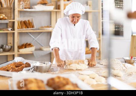 Reife Bäckerin, die Teig mit einer Nadel in einer Bäckerei rollt Stockfoto