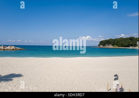 Shirarahama, Japan - 25. Oktober 2023: Ein weiträumiger weißer Sandstrand mit türkisfarbenem Wasser, einem felsigen Wellenbrecher auf der linken Seite und einer bewaldeten Landzunge mit ein paar Gebäuden auf der rechten Seite unter einem klaren blauen Himmel. Stockfoto