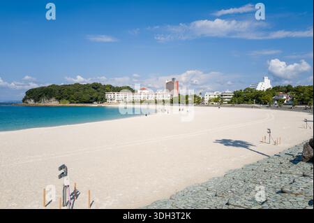 Shirarahama, Japan - 25. Oktober 2023: Ein breiter Sandstrand mit verstreuten Menschen, die in der Nähe der Küste spazieren, neben türkisfarbenem Wasser, mit modernen Gebäuden, Hotels und einem bewaldeten Bereich, der im Hintergrund unter einem klaren blauen Himmel zu sehen ist. Stockfoto