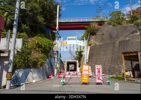 Shirarahama, Japan - 25. Oktober 2023: Eine Straße ist durch Verkehrsbarrieren und japanische Straßenschilder in der Nähe einer Unterführung blockiert, mit einer roten Brücke und Überlandleitungen, die über und an den Hängen sichtbar sind. Stockfoto