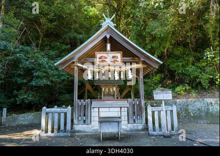 Shirarahama, Japan - 25. Oktober 2023: Ein kleines Shinto-Schrein-Gebäude mit Holzdach, Seil mit Papierschlangen und zwei traditionellen Maskendekorationen über dem Eingang, umgeben von dichtem Wald und Steinzäunen. Stockfoto