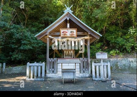 Shirarahama, Japan - 25. Oktober 2023: Ein kleines Shinto-Schrein-Gebäude mit Holzdach, Seil mit Papierschlangen und zwei traditionellen Maskendekorationen über dem Eingang, umgeben von dichtem Wald und Steinzäunen. Stockfoto