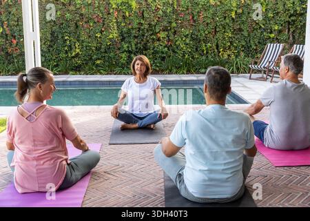 Verschiedene Freunde, die einer älteren Frau beim Üben folgten, saßen auf Yogamatten auf der Backsteinterrasse neben dem Pool Stockfoto