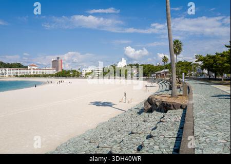 Shirarahama, Japan - 25. Oktober 2023: Ein breiter Sandstrand mit verstreuten Menschen, die in der Nähe der Küste spazieren, neben einem Steinweg, der von Palmen gesäumt ist, und im Hintergrund sichtbare moderne Gebäude unter einem klaren blauen Himmel. Stockfoto