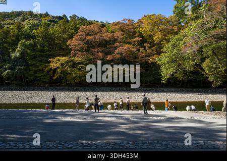 ISE, Japan - 29. Oktober 2023: Gruppen von Menschen stehen und sitzen am Rand des Mitarashiba Reinigungsbeckens am Fluss, mit Bäumen, die Herbstlaub im Hintergrund zeigen. Stockfoto