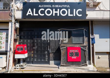 Shirarahama, Japan - 25. Oktober 2023: An der Außenseite einer Bar befindet sich ein großes Schild mit der Aufschrift „ALCOHOL IS BEAUTIFUL ALCOHOL“ über dem Eingang, mit zusätzlichen japanischen Schildern und einer geschlossenen schwarzen Tür bei Tageslicht. Stockfoto
