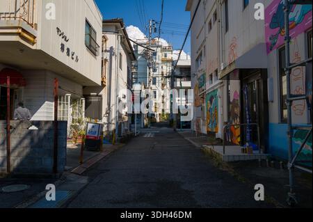 Shirarahama, Japan - 25. Oktober 2023: Eine enge Straße, die von flachen Gebäuden, farbenfrohen Wandmalereien, japanischen Schildern, Strommasten und Freileitungen gesäumt ist. Stockfoto