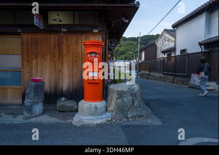 Hiragawa, Japan - 24. Oktober 2023: An einer Straßenecke neben einem traditionellen Holzgebäude steht ein roter zylindrischer Postkasten, an dem eine Person vorbeiläuft und Wohnhäuser im Hintergrund sichtbar sind. Stockfoto