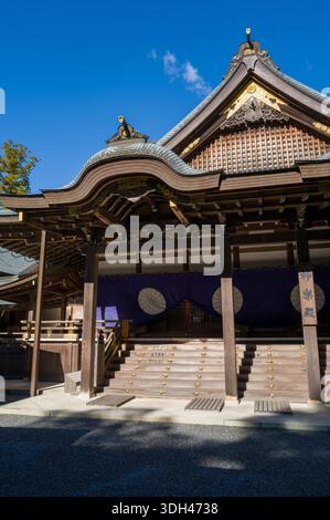 ISE, Japan - 29. Oktober 2023: Die Eingangstreppe und das verzierte Dach der Kaguraden Hall in Ise Jingu werden mit traditioneller Holzarchitektur, goldenen dekorativen Elementen und lila Vorhängen mit weißen Emblemen unter einem klaren blauen Himmel gezeigt. Stockfoto