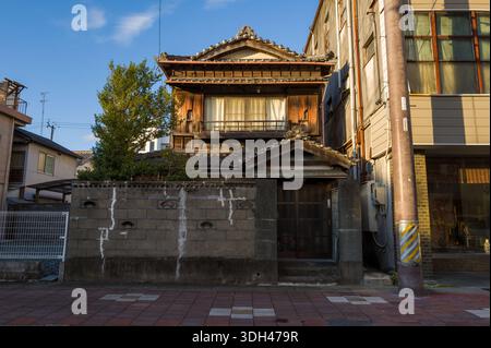 ISE, Japan - 28. Oktober 2023: Ein traditionelles zweistöckiges Holzhaus mit einem gekachelten Dach und einer Betonmauer ist auf einer Stadtstraße zu sehen, mit angrenzenden modernen Gebäuden und einem Baum, der bei klarem Tageslicht sichtbar ist. Stockfoto