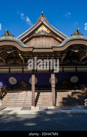ISE, Japan - 29. Oktober 2023: Das kunstvoll geschwungene Dach und die Eingangstreppe der Kaguraden Hall in Ise Jingu werden mit traditioneller Holzarchitektur, goldenen dekorativen Elementen und lila Vorhängen mit weißen Emblemen unter einem klaren blauen Himmel gezeigt. Stockfoto