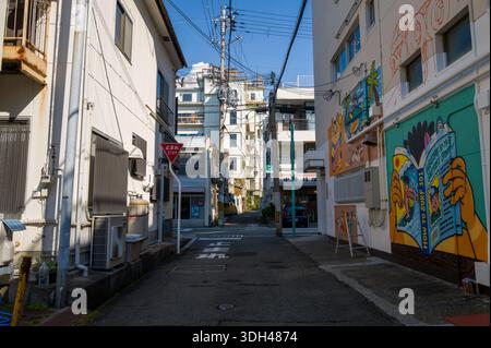 Shirarahama, Japan - 25. Oktober 2023: Eine enge Stadtgasse zeigt bunte Wandmalereien am rechten Gebäude, ein japanisches Stoppschild, Strommasten mit Freileitungen und flache Wohn- und Geschäftsgebäude unter klarem blauen Himmel. Stockfoto