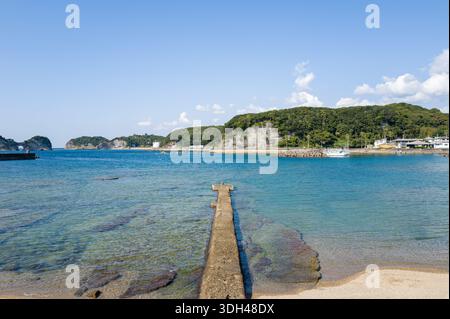 Shirarahama, Japan - 25. Oktober 2023: Ein Betonpier erstreckt sich im Hafen von Shirarahama in klares blaues Wasser, mit bewaldeten Hügeln, felsiger Küste und mehreren Booten und Gebäuden, die im Hintergrund unter klarem Himmel zu sehen sind. Stockfoto