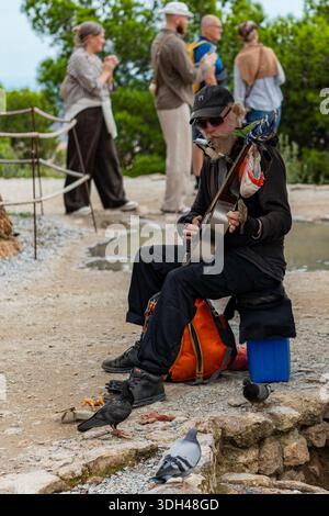 Barcelona, Spanien - 20. Oktober 2025: Ein bärtiger Straßenmusiker spielt eine Resonatorgitarre und Mundharmonika, umgeben von Tauben im Park Güell. Stockfoto