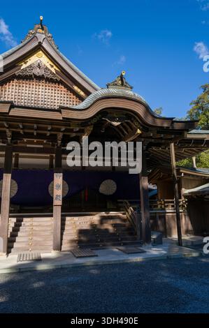 ISE, Japan - 29. Oktober 2023: Das kunstvoll verzierte Holzdach und die Eingangsstufen der Kaguraden Hall in Ise Jingu sind mit goldenen dekorativen Elementen, violetten Vorhängen mit weißen Emblemen und traditionellen architektonischen Details unter einem klaren blauen Himmel zu sehen. Stockfoto
