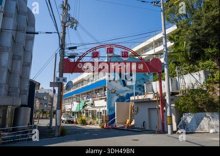 Shirarahama, Japan - 25. Oktober 2023: Ein roter Bogen mit japanischem Text markiert den Eingang zu einer Einkaufsstraße mit Freileitungen, nahe gelegenen Gebäuden und einem Wandbild mit Meerestieren, das im Hintergrund sichtbar ist. Stockfoto