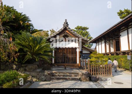 Nara, Japan - 4. November 2023: Das Bild zeigt ein kleines traditionelles Holzgebäude mit einem gekachelten Dach am Horyu-JI-Tempel, umgeben von üppigen Pflanzen und Steintreppen, wobei zwei Personen in der Nähe laufen. Stockfoto