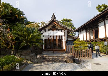 Nara, Japan - 4. November 2023: Das Bild zeigt ein kleines traditionelles Holzgebäude mit einem gekachelten Dach am Horyu-JI-Tempel, umgeben von üppigen Pflanzen und Steintreppen, in der Nähe mehrere Personen laufen. Stockfoto