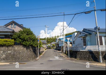 Shirarahama, Japan - 25. Oktober 2023: Eine ruhige Wohnstraße, gesäumt von Einfamilienhäusern, Steinmauern, Versorgungsmasten und Freileitungen unter einem klaren blauen Himmel mit großen Wolken im Hintergrund. Stockfoto
