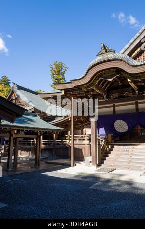 ISE, Japan - 29. Oktober 2023: Der Innenhof und der Eingangsbereich der Kaguraden Hall in Ise Jingu sind mit traditioneller Holzarchitektur, einem kunstvoll geschwungenen Dach, goldenen dekorativen Elementen und lila Vorhängen mit weißem Emblem unter einem klaren blauen Himmel sichtbar. Stockfoto