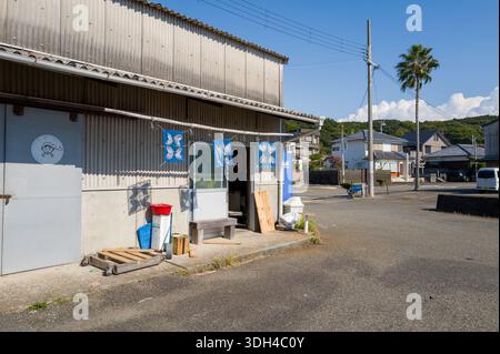 Shirarahama, Japan - 25. Oktober 2023: Das Äußere eines kleinen Ladens in der Nähe des Hafens mit blauen Bannern, einer Metalltür mit Fischlogo, Mülltonnen, Holzpaletten und einer Bank mit Wohnhäusern, eine Palme und Stromleitungen, die im Hintergrund unter klarem Himmel sichtbar sind. Stockfoto