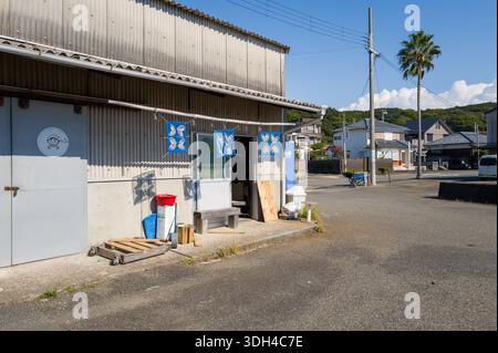 Shirarahama, Japan - 25. Oktober 2023: Das Äußere eines kleinen Ladens in der Nähe des Hafens mit blauen Bannern, einer Metalltür mit einem Fischlogo, Mülltonnen und Holzpaletten, mit Wohnhäusern und einer Palme, die im Hintergrund unter klarem Himmel zu sehen ist. Stockfoto