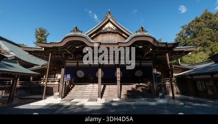 ISE, Japan - 29. Oktober 2023: Die Vorderansicht der Kaguraden Hall in Ise Jingu zeigt traditionelle Holzarchitektur, ein kunstvoll geschwungenes Dach mit goldenen dekorativen Elementen und lila Vorhänge mit weißen Emblemen unter einem klaren blauen Himmel. Stockfoto