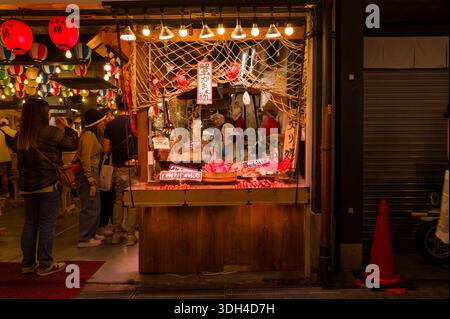 Kyoto, Japan - 19. Oktober 2023: Das Bild zeigt einen hell beleuchteten Imbissstand in der Nacht im Zentrum von Kyoto, mit Meeresfrüchten, japanischen Schildern, roten Laternen und mehreren Personen, die vor dem Theken stehen. Stockfoto