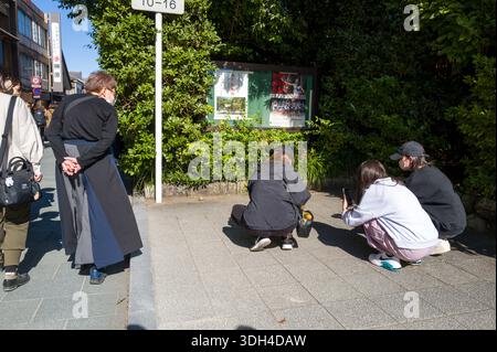 ISE, Japan - 29. Oktober 2023: Drei Personen hocken sich auf einem Bürgersteig und machen Fotos von einer Katze in der Nähe von Grün und einer Informationstafel, während andere Fußgänger an einem sonnigen Tag vorbeilaufen. Stockfoto