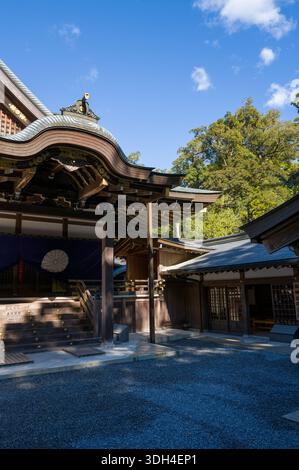 ISE, Japan - 29. Oktober 2023: Das kunstvoll verzierte Holzdach und der Innenhof der Kaguraden Hall in Ise Jingu sind mit traditionellen architektonischen Details, goldenen dekorativen Elementen und lila Vorhängen mit weißem Emblem unter einem klaren blauen Himmel zu sehen. Stockfoto