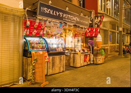 Kyoto, Japan - 19. Oktober 2023: Das Bild zeigt den Essensstand Sally's Kitchen am Nishiki Market in Kyoto bei Nacht mit beleuchteten Vitrinen, farbenfrohen Schildern für Desserts und Eis und einem großen Plastikeiskegel-Modell außerhalb des Ladens. Stockfoto