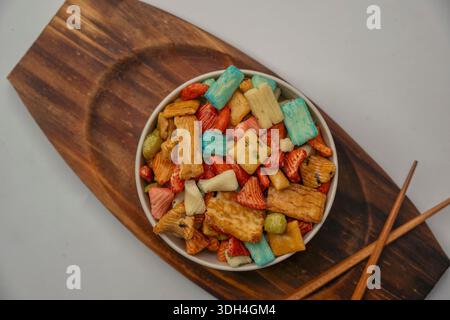 Eine Schüssel mit verschiedenen japanischen Crackern steht auf einem Holztablett. Das Fach wird auf einem weißen Hintergrund platziert. Die Snacks sind in verschiedenen Farben und Formen erhältlich Stockfoto