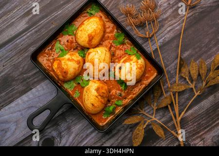 Eine schwarze Pfanne mit Eiern Masala / Eiercurry auf einem Holztisch. Die Eier werden in einer Reihe angeordnet und die Soße wird über die Pfanne verteilt. Die Szene hat ein W Stockfoto