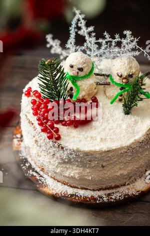Eine festliche weiße Kokosnussschicht-Torte, dekoriert mit essbaren Schneemännern und roten Johannisbeeren, mit einer Scheibe auf einem Teller, die eine dunkle Beerenkonfitüre enthüllt. Stockfoto