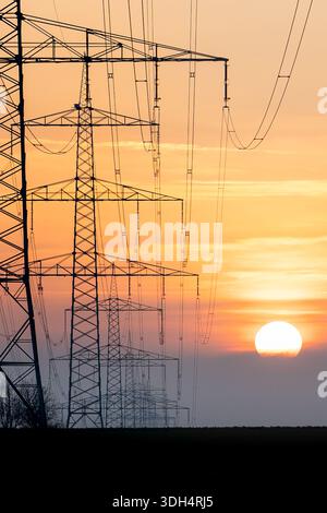 Hochspannungs-Übertragungstürme und Stromleitungen, die sich vor dem Sonnenunterganghimmel im ländlichen Deutschland als Symbol für Energieversorgung, Infrastruktur und Elektro darstellen Stockfoto