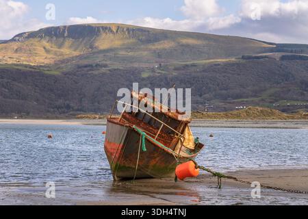 Barmouth, Wales, 6. März 2024: Verlassenes und verwittertes Fischerboot kippte bei Ebbe am Ufer. Verrosteter Rumpf, Festmacherkette und Boje im Vorderteil Stockfoto