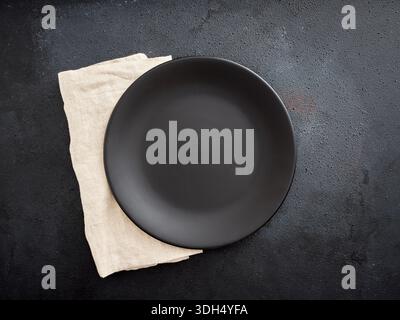 Leere schwarze Platte auf einer beigen Leinenserviette, die einen minimalistischen dunklen Hintergrund bietet und die Perspektive von oben bietet Stockfoto