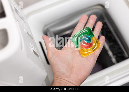 Mann, der Waschmittelkapsel drinnen in die Waschmaschine steckt, Nahaufnahme. Buntes Waschgel in Kapsel. Wäschetücher waschen. Das Konzept von war Stockfoto
